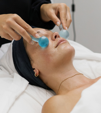 A person lies on a treatment bed with eyes closed while another uses blue glass facial globes to massage their face—a relaxing spa treatment, perfect for those seeking rejuvenating things to do in Costa Mesa.