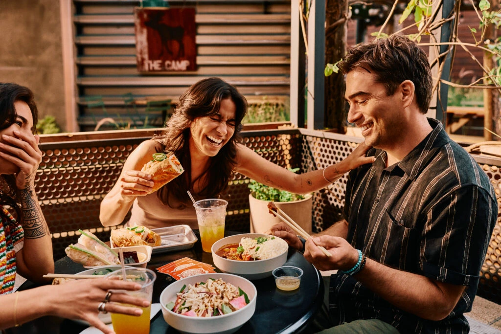 Three friends sit at an outdoor table, laughing and enjoying tacos, salads, and drinks. One woman covers her mouth while laughing, another holds a taco, and a man looks at his phone, smiling.