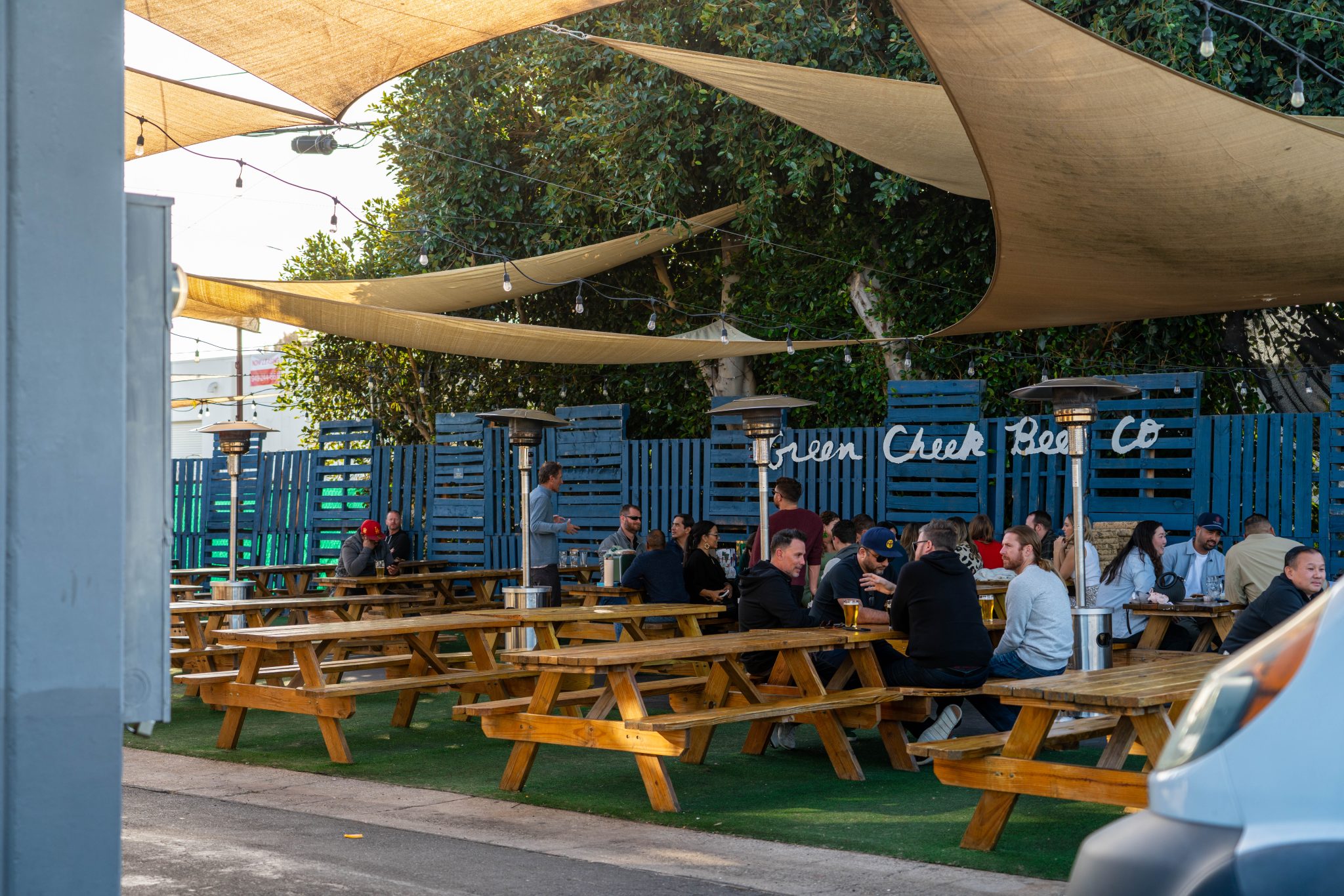 People sit at wooden picnic tables under shade sails in an outdoor seating area at a brewery. The area has string lights, heaters, blue fence walls, and the sign Green Creek Beer Co in the background.