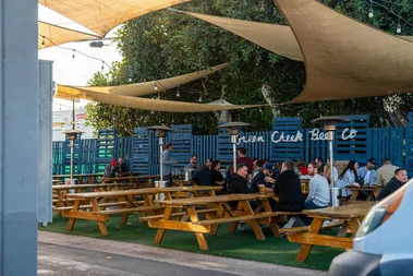 People sit at wooden picnic tables in an outdoor seating area under tan shade sails. The space has string lights, patio heaters, and a blue fence with the words “Green Cheek Beer Co” in the background.