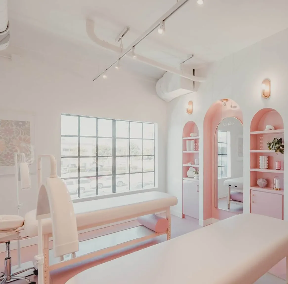 A bright, modern beauty treatment room with pastel pink and white decor, two treatment beds, large windows, built-in shelves with supplies, and arched mirrors—a perfect spot to unwind among the top things to do in Costa Mesa.