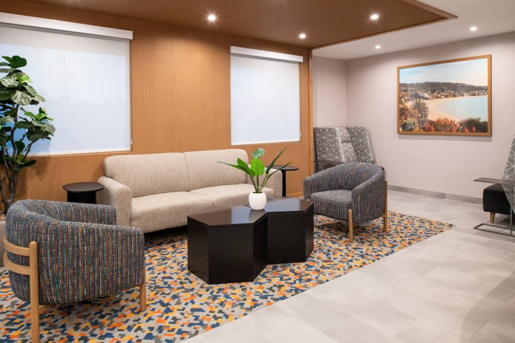 Modern lounge area with a beige sofa, two patterned armchairs, geometric rug, black hexagonal coffee tables, and lush plants. Ideal for gatherings or as inspiration for event venues in Costa Mesa. Large windows flood the space with natural light.