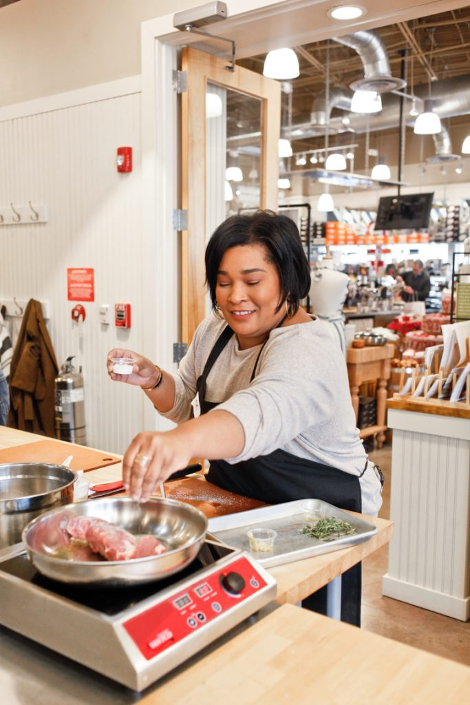 A woman cooking in a kitchen store, smiling as she seasons meat in a frying pan on an electric stove—one of the fun things to do in Costa Mesa—with shelves and shoppers visible in the background.