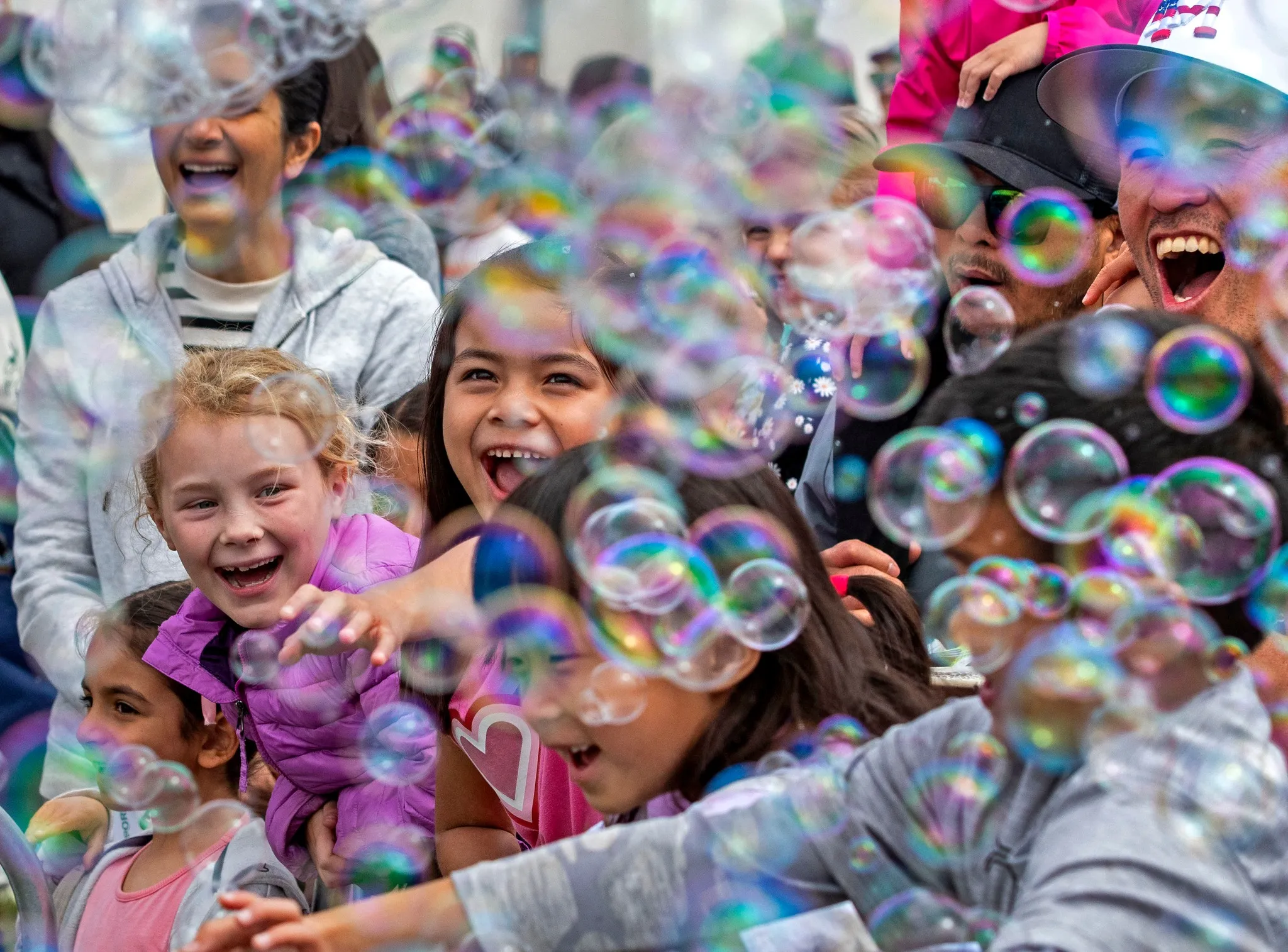 A group of children and adults laugh and reach out excitedly as colorful bubbles float around them at a lively outdoor event in Costa Mesa. The joyful scene reflects the playful spirit of annual events Costa Mesa is known for.