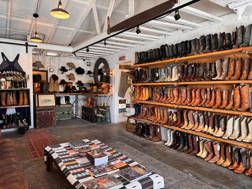 A stylish store interior with white ceilings, shelves filled with various cowboy boots, some hats and accessories on display, and a table covered in books and magazines on a patterned rug.