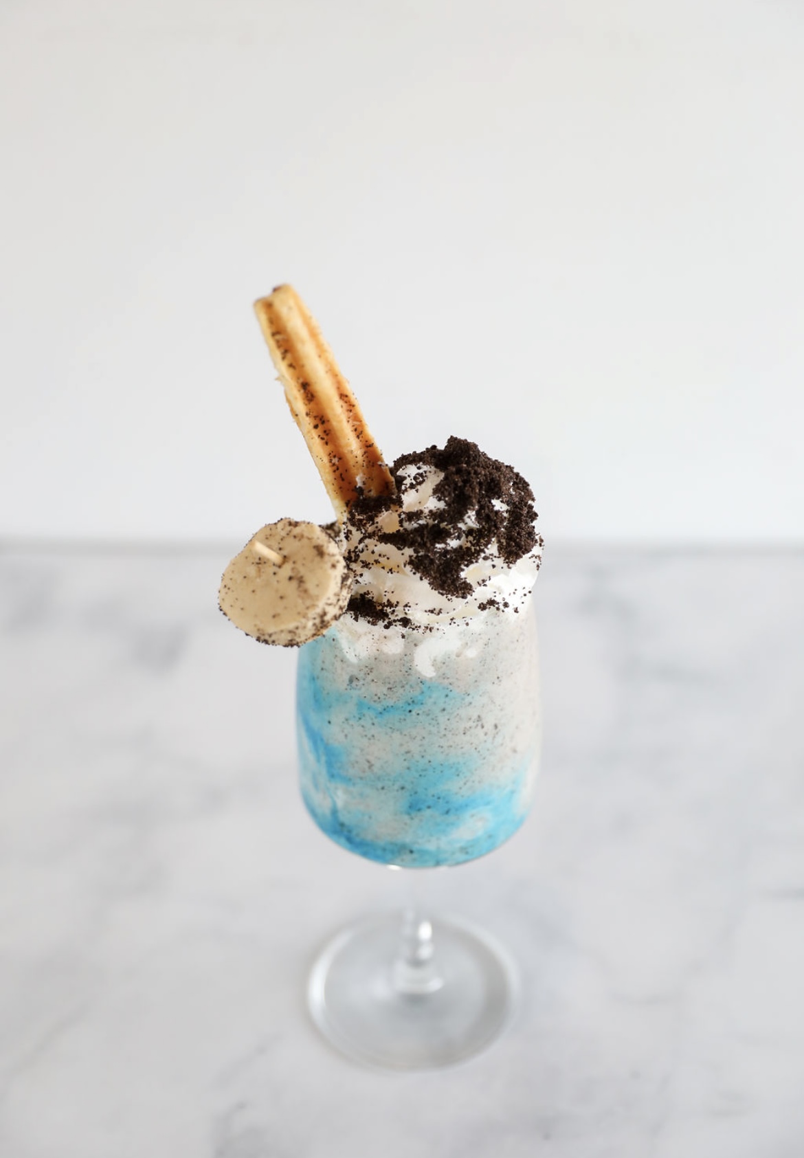 A tall glass of a blue and white frozen drink topped with whipped cream, cookie crumbs, a round cookie, and a toasted wafer on a marble surface.