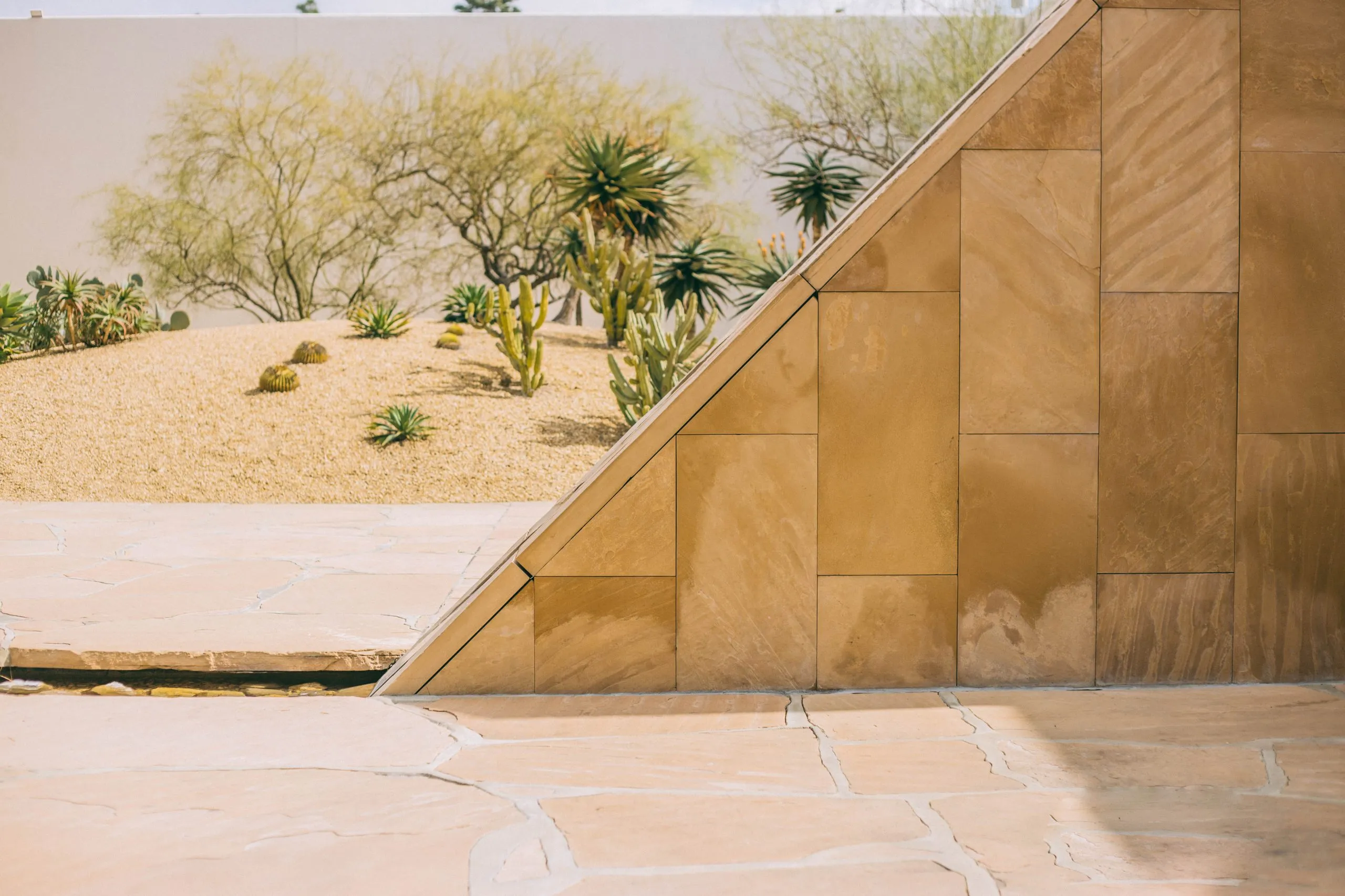 A geometric structure with tan stone tiles sits beside a desert garden featuring sand, rocks, and various cacti and succulents, with a white wall and sparse trees in the background.