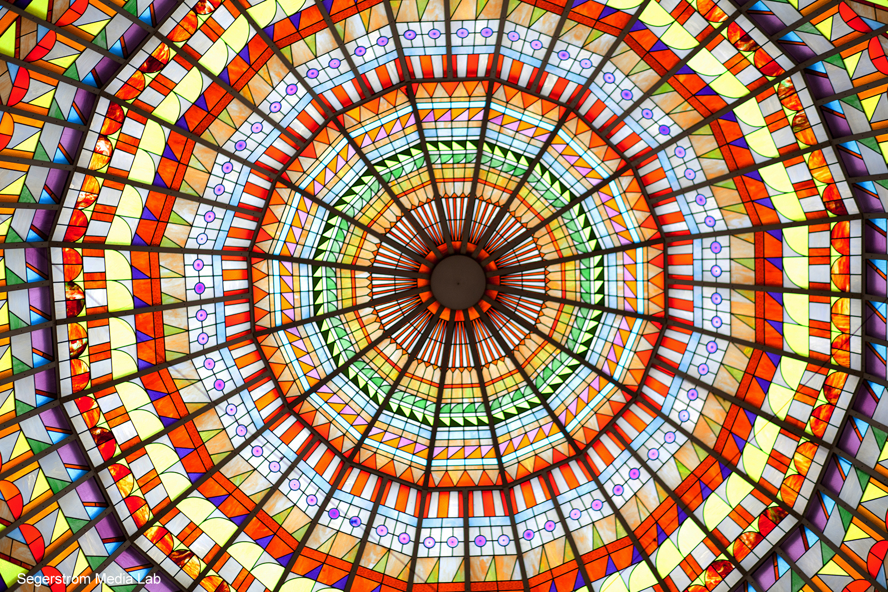 A vibrant stained glass dome ceiling with intricate geometric patterns in bright orange, yellow, red, green, and purple radiates from a central point—perfect for capturing the creative spirit of the Costa Mesa Art Walk.
