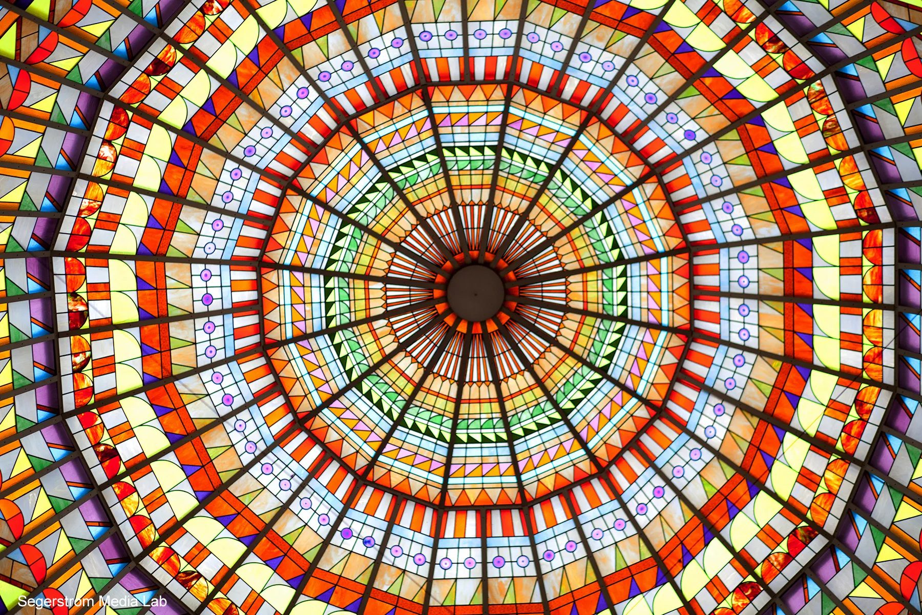 A vibrant stained glass dome ceiling with intricate geometric patterns in bright orange, yellow, red, green, and purple radiates from a central point—perfect for capturing the creative spirit of the Costa Mesa Art Walk.