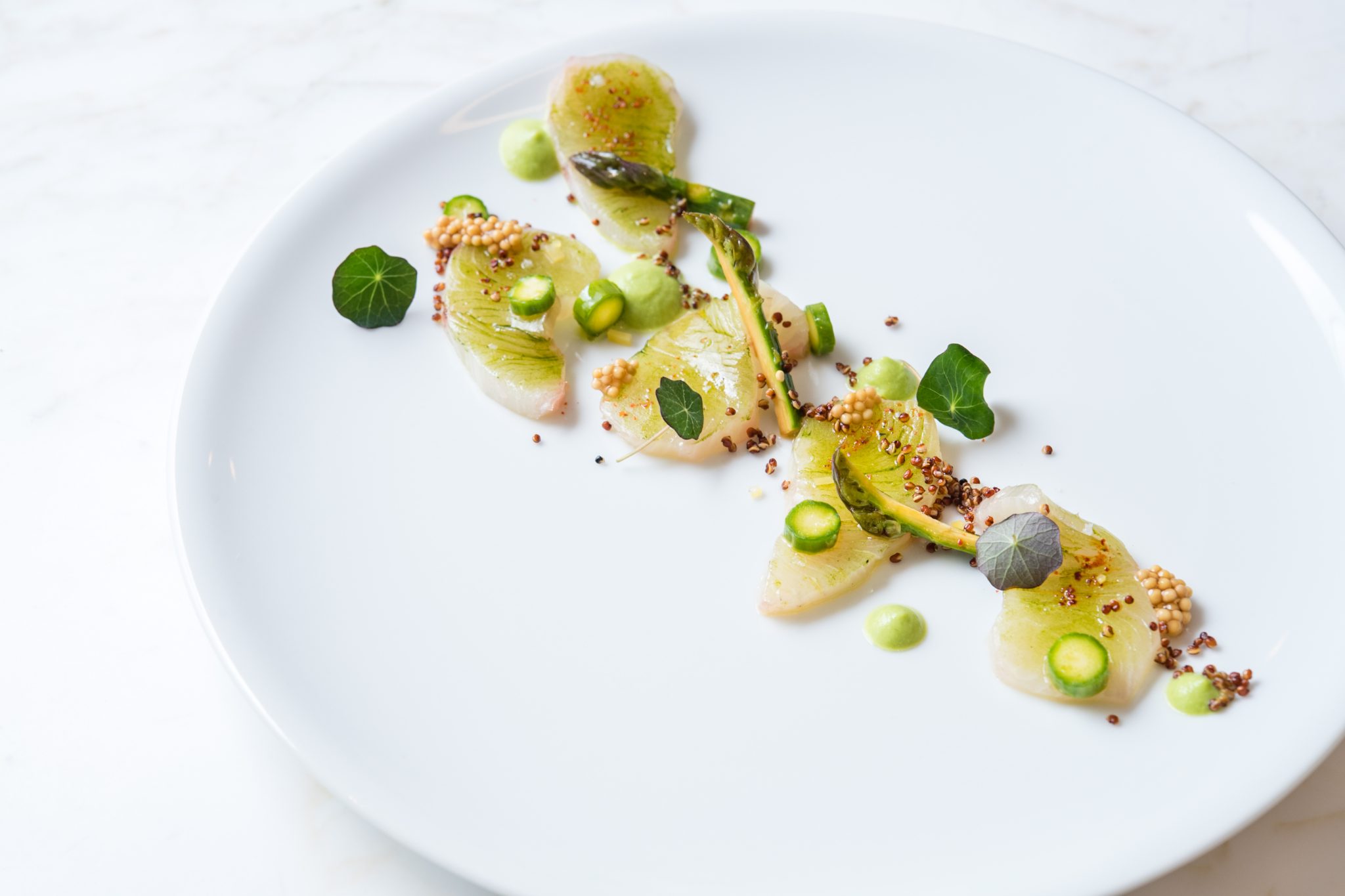 A white plate with artfully arranged slices of fish, green asparagus, small leaves, mustard seeds, and green sauce dots on a white background. The presentation is minimalist and elegant.