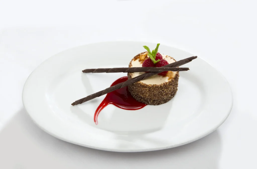A round cheesecake garnished with two chocolate sticks, a raspberry, and a mint sprig, sits on a white plate with a swirl of red berry sauce.