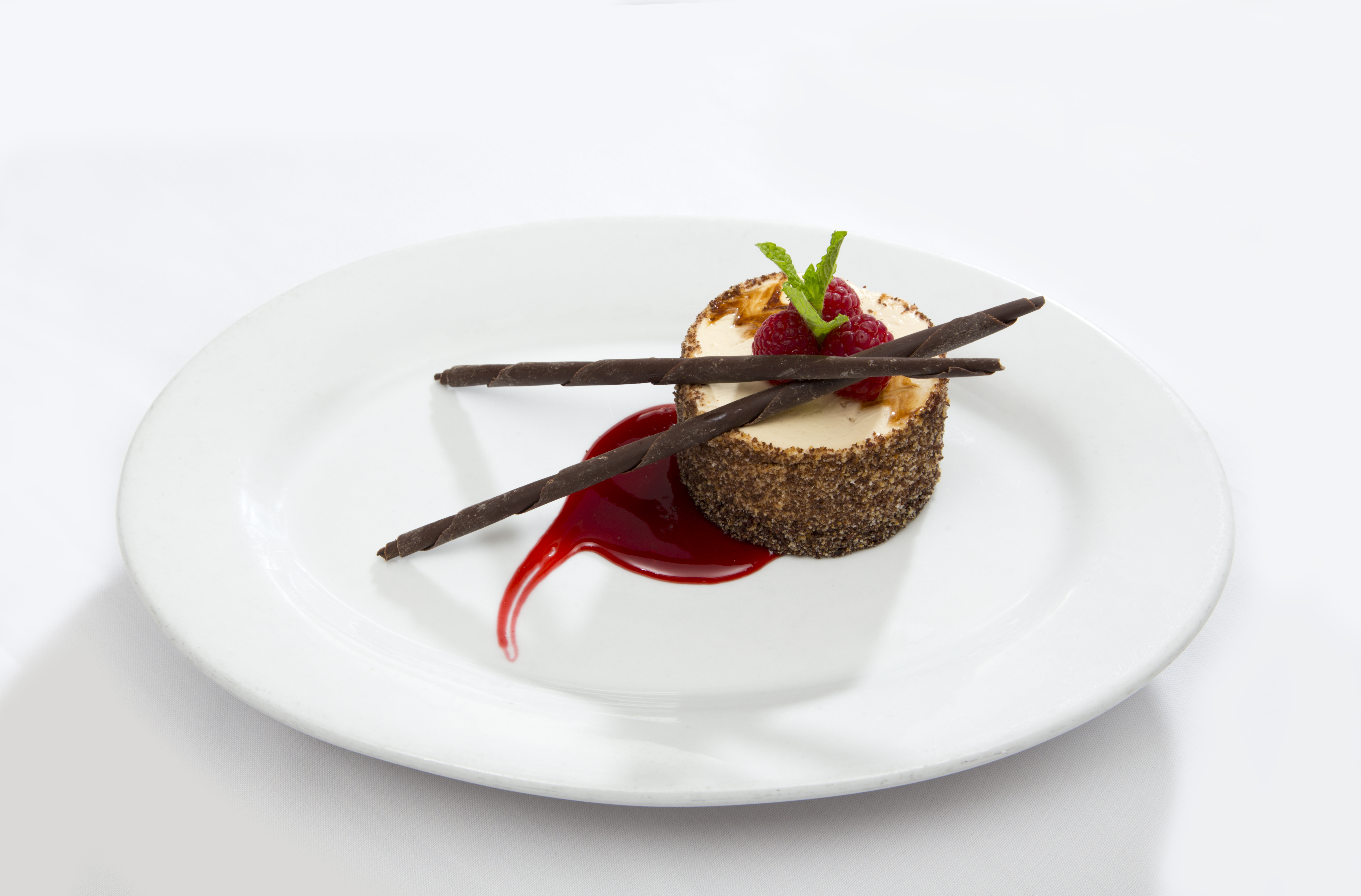 A round cheesecake garnished with two chocolate sticks, a raspberry, and a mint sprig, sits on a white plate with a swirl of red berry sauce.