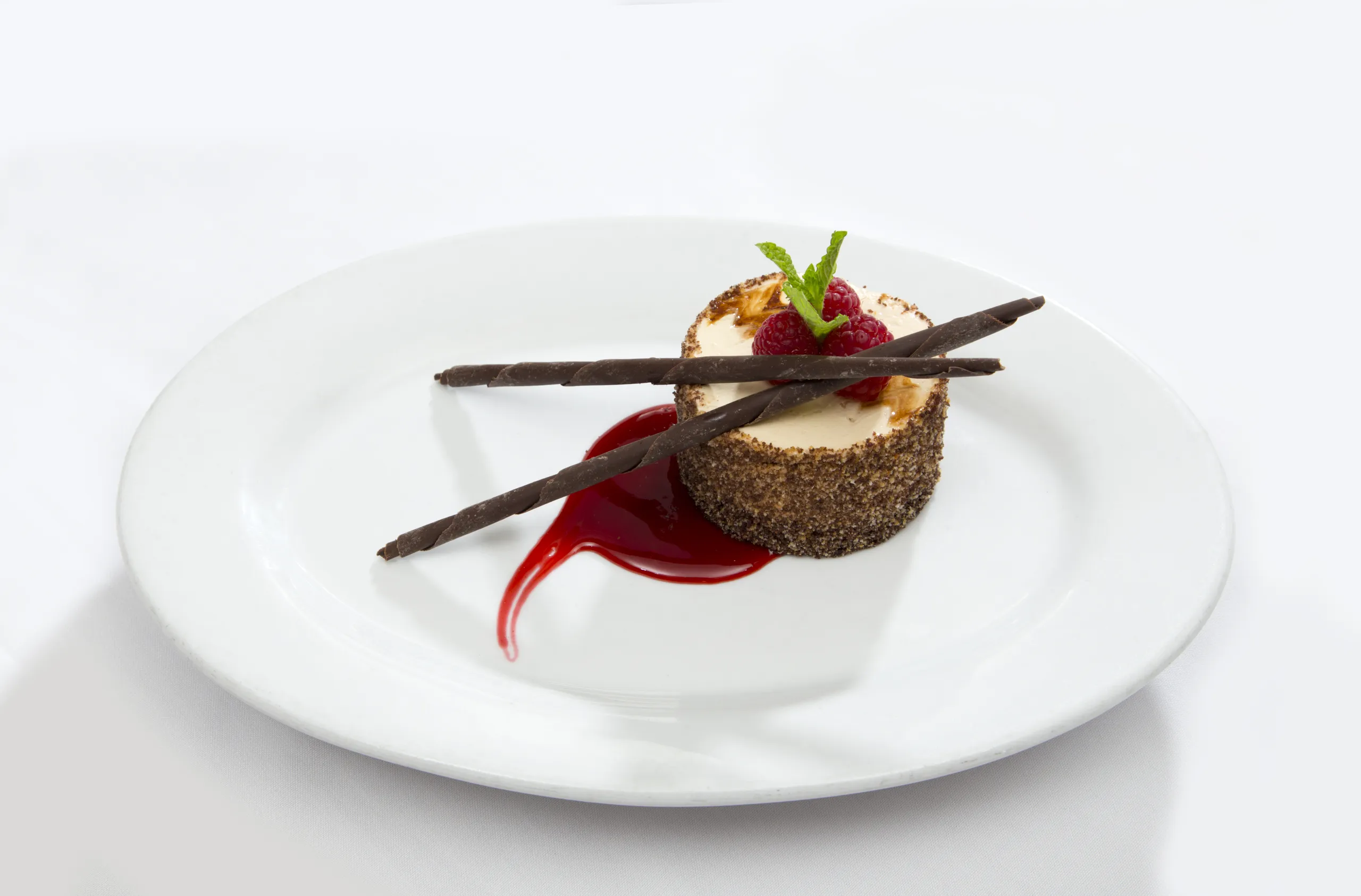 A round cheesecake garnished with two chocolate sticks, a raspberry, and a mint sprig, sits on a white plate with a swirl of red berry sauce.