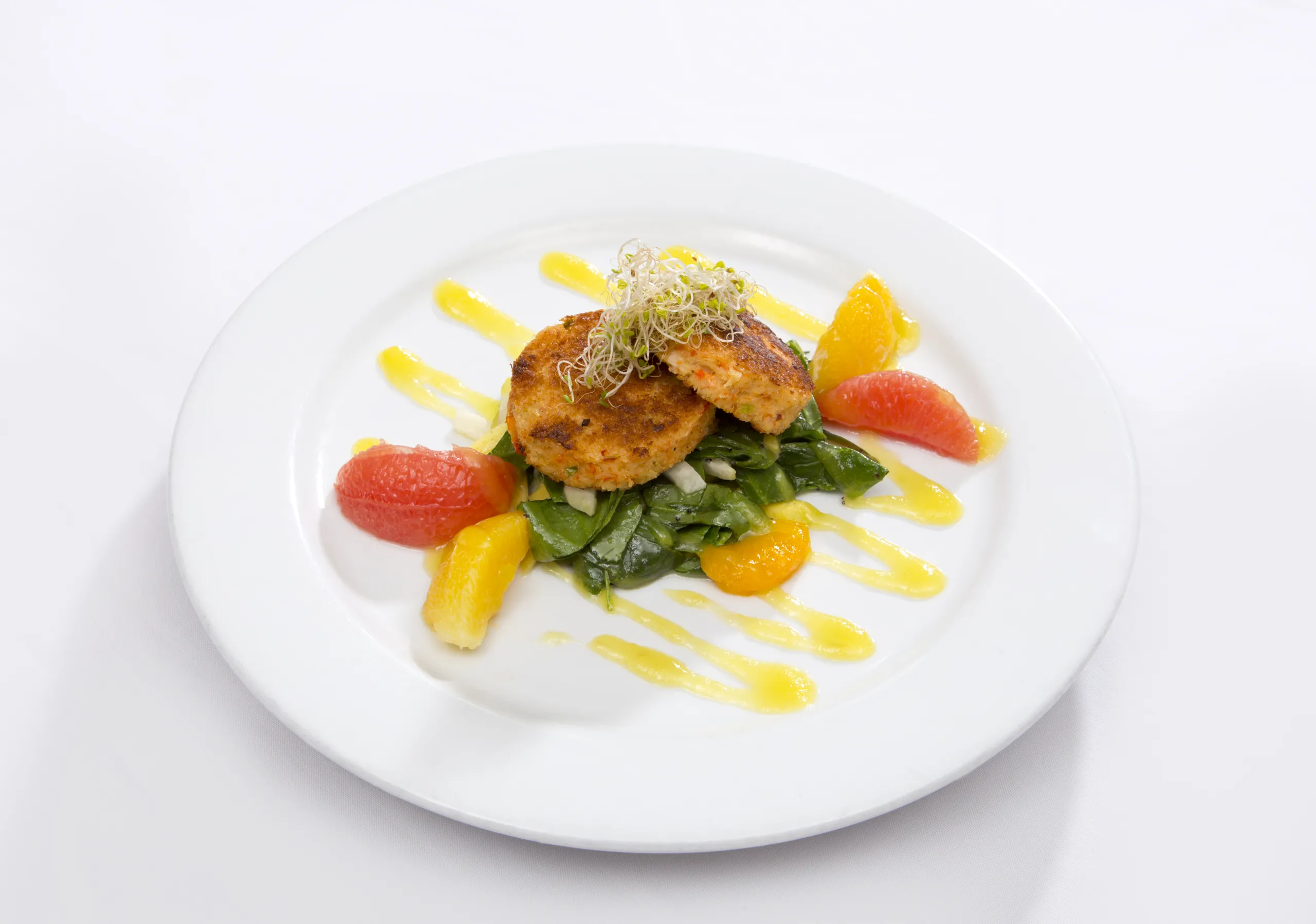 Two golden-brown patties on a bed of spinach, topped with sprouts, served with orange and grapefruit segments, and drizzled with yellow sauce on a white plate.