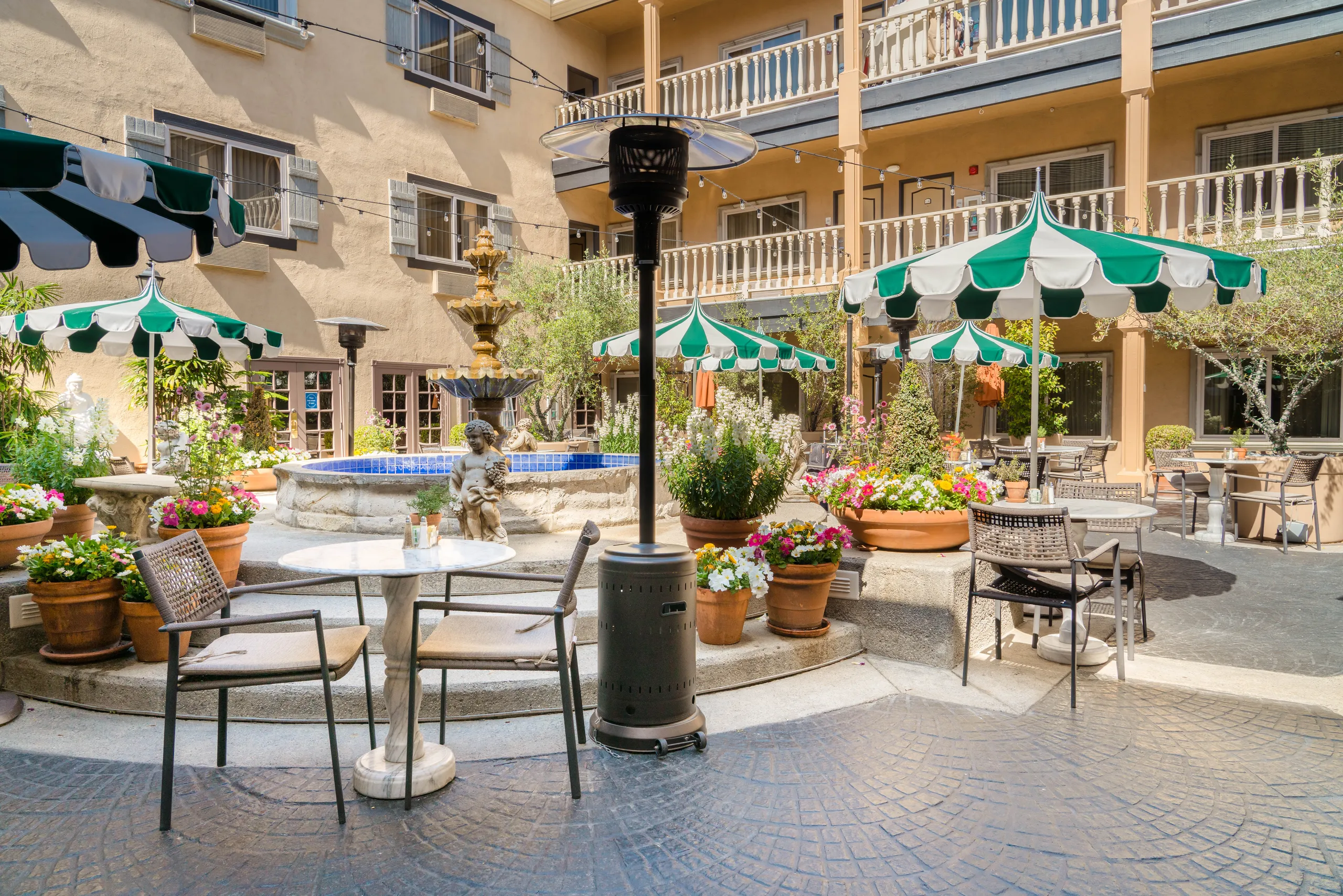 Sunny courtyard with a central fountain, green-and-white striped umbrellas, outdoor tables and chairs, potted flowers, and a two-story beige building—it's the perfect spot for the best brunch Costa Mesa has to offer.
