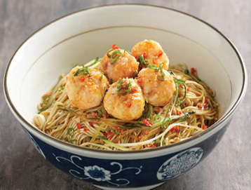 A bowl of noodles topped with five golden shrimp balls, chopped green onions, and red chili pieces, served in a blue and white patterned bowl.