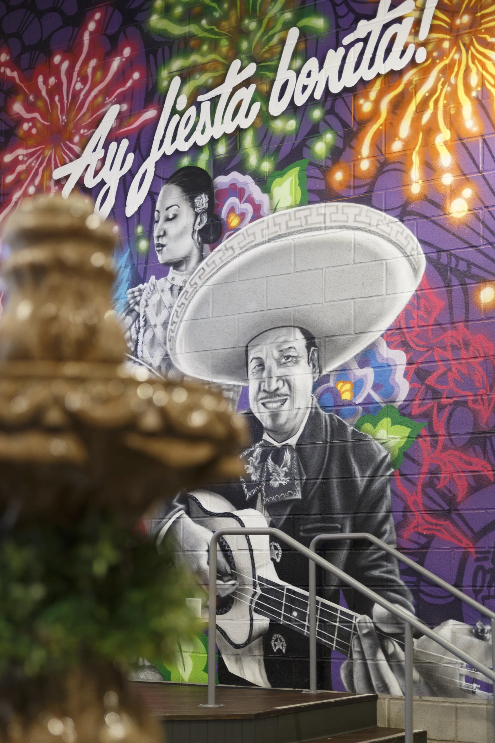 A colorful mural features a man in a charro outfit playing guitar and a woman in traditional dress, with festive fireworks and Ay, fiesta bonita! written above. A decorative fountain is blurred in the foreground.