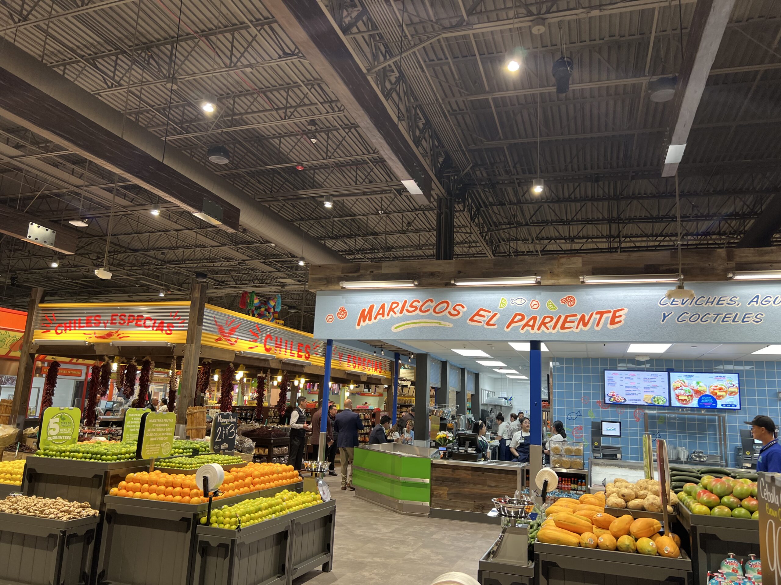 A vibrant grocery store interior with fresh produce displays in the foreground and neon signs reading Mariscos El Pariente and Chiles, Especias above seafood and spice sections. Shoppers and staff are visible.