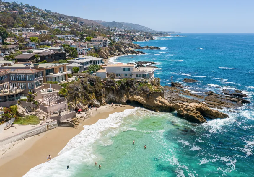Aerial view of a coastal neighborhood with modern houses on cliffs above a sandy beach and turquoise ocean. People are swimming and relaxing, while hills and more homes are visible—just miles from future FIFA World Cup 2026 Los Angeles festivities.