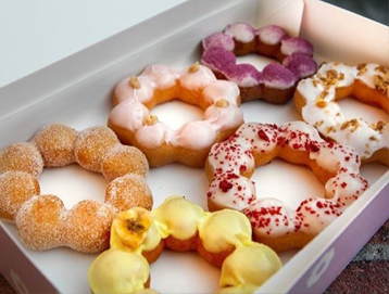 A box of six assorted mochi donuts, each with a unique flavor and colorful toppings, arranged neatly in white packaging. The donuts have a distinctive ring shape made of round, connected segments.