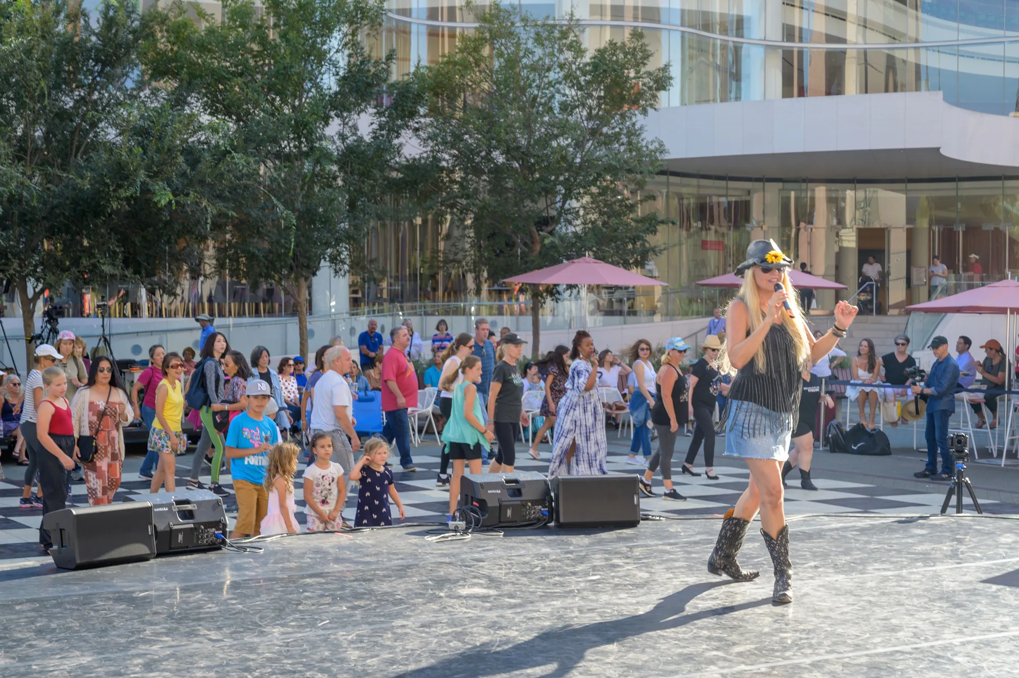 A woman in a hat and boots performs on an outdoor stage as a diverse crowd of people, including children, watch and gather in a sunny plaza with modern buildings and trees in the background.