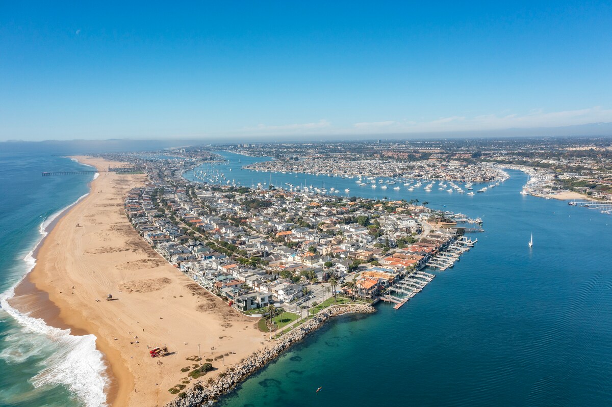 Aerial view of a long, narrow peninsula with sandy beaches on one side and a marina with boats on the other, lined with houses and surrounded by blue ocean and clear skies.