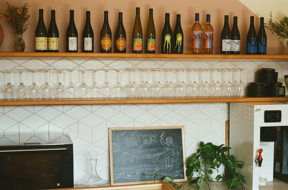 A kitchen shelf displays colorful wine bottles above hanging glasses. Below, a chalkboard with drawings sits on the counter, surrounded by a plant, microwave, and coffee machine—perfect inspiration for things to do in Costa Mesa.