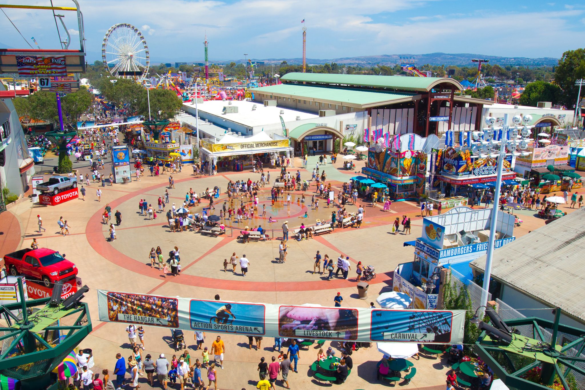 A lively outdoor fairground scene with crowds of people walking, colorful booths, amusement rides including a Ferris wheel, banners, and festive decorations under a bright blue sky.