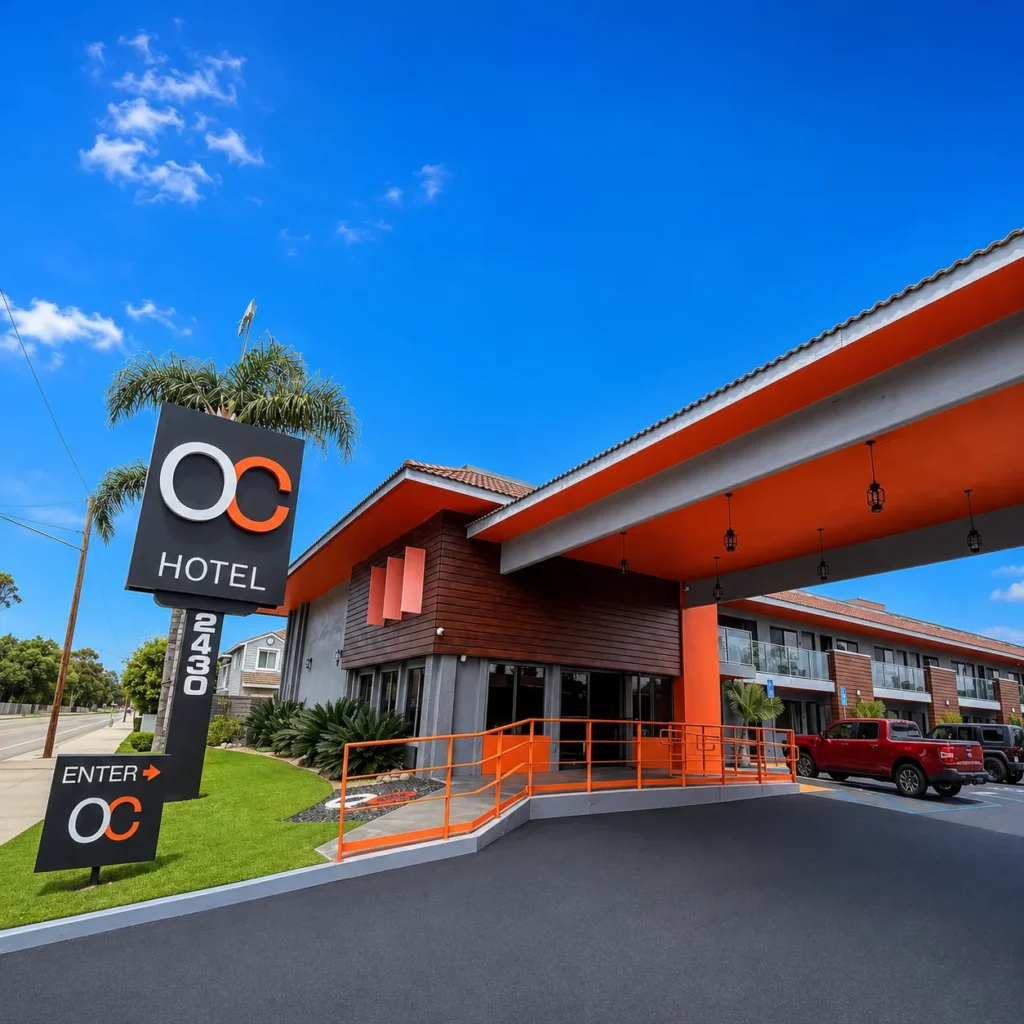 A modern hotel exterior with a large sign reading OC HOTEL and ENTER OC 2430. This standout among hotels Costa Mesa features orange accents, a covered entrance, palm trees, and parked vehicles under a clear blue sky.