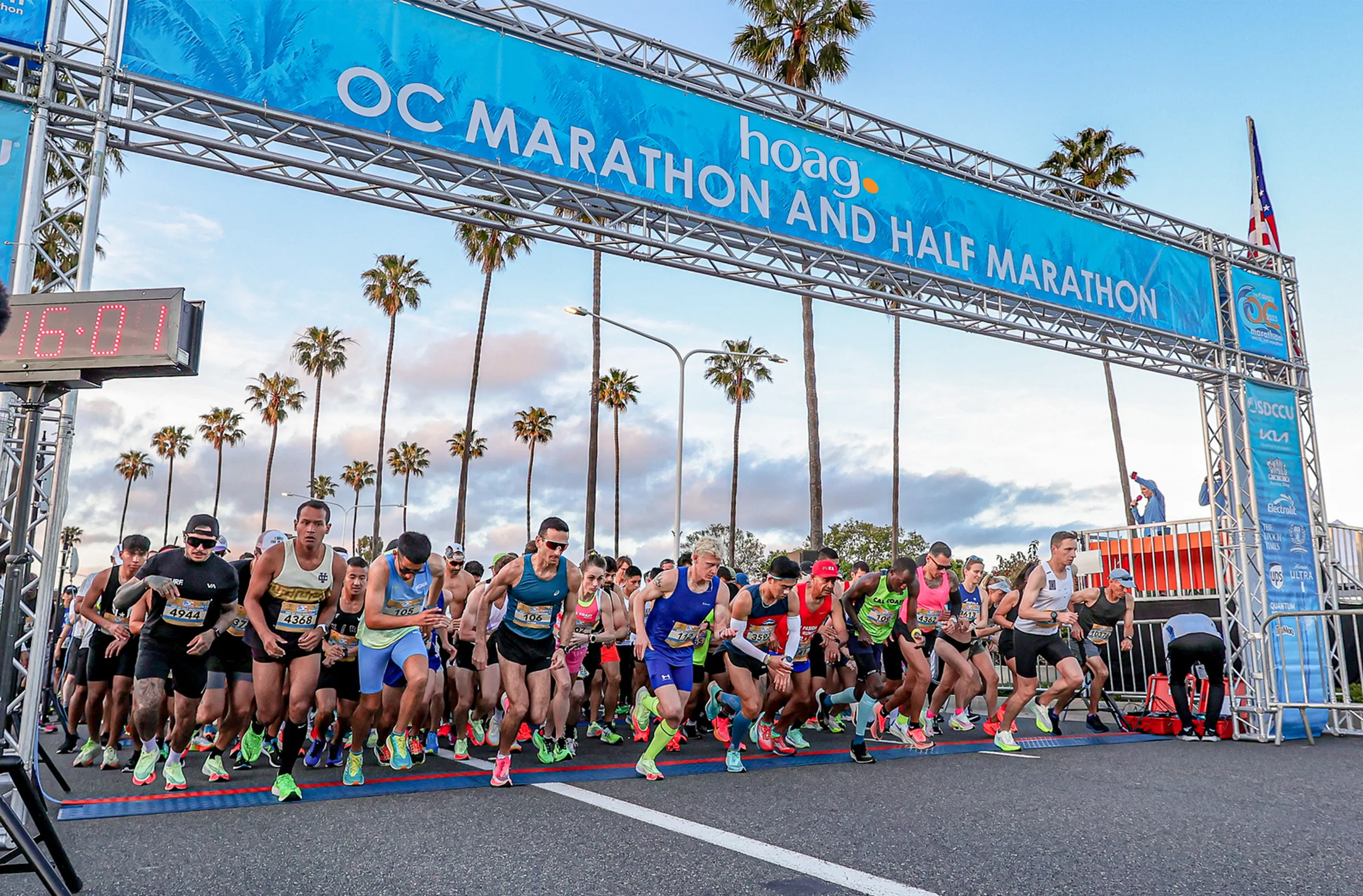 Runners in athletic gear begin a race at the starting line of the OC Marathon and Half Marathon, one of the top annual events Costa Mesa hosts, beneath a blue banner, with palm trees and a partly cloudy sky in the background.