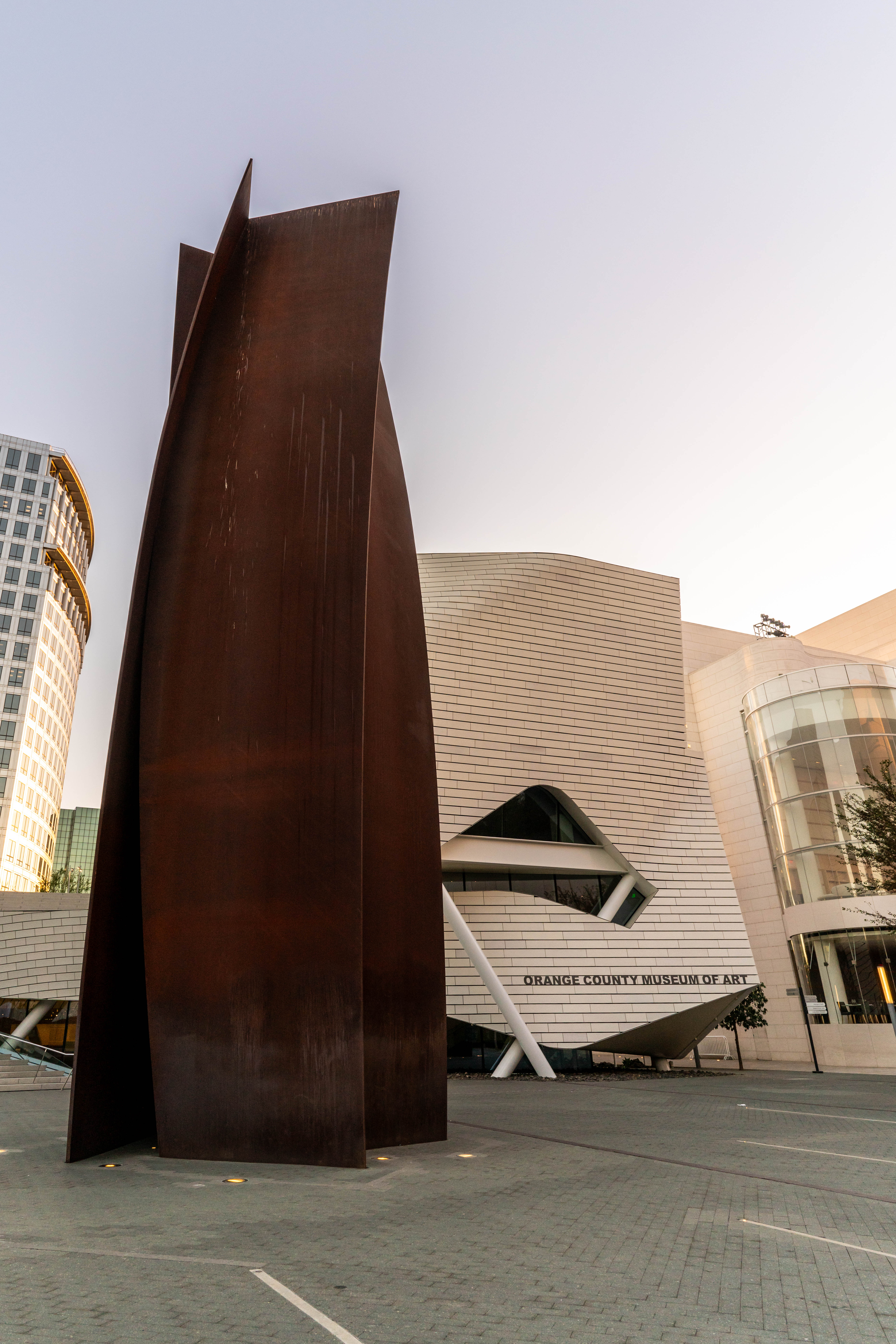 A tall, abstract rust-colored metal sculpture stands in front of the modern facade of the Orange County Museum of Art, a highlight along the Costa Mesa Art Walk, with curved white walls and glass windows in the background.