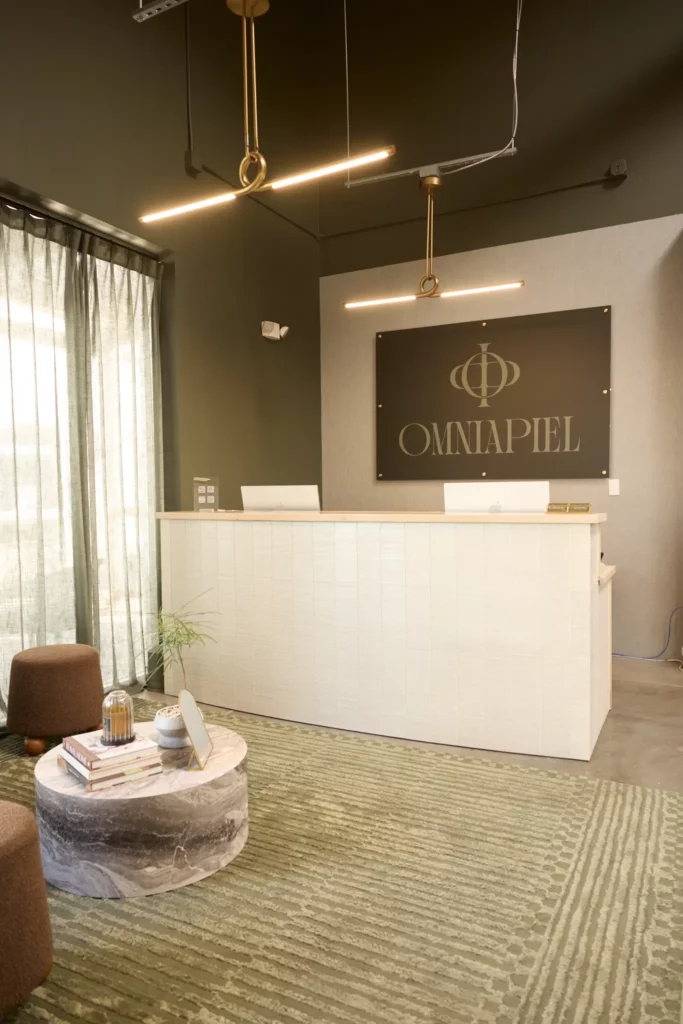 A modern reception area with a white front desk, two laptops, a sign reading OMNIAPIEL on the wall, and decor that’s inspired by things to do in Costa Mesa—brown chairs, green rug, marble coffee table with books, and hanging gold lights.