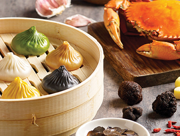 A bamboo steamer holds five colorful soup dumplings; beside it are a cooked crab, black truffles, garlic cloves, and a small bowl of truffle slices, all arranged on a wooden table.