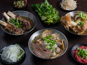 Bowls of Vietnamese pho with assorted meats, herbs, lime wedges, bean sprouts, sliced onions, and raw beef, arranged on a dark table. The dishes are garnished with green onions and herbs.