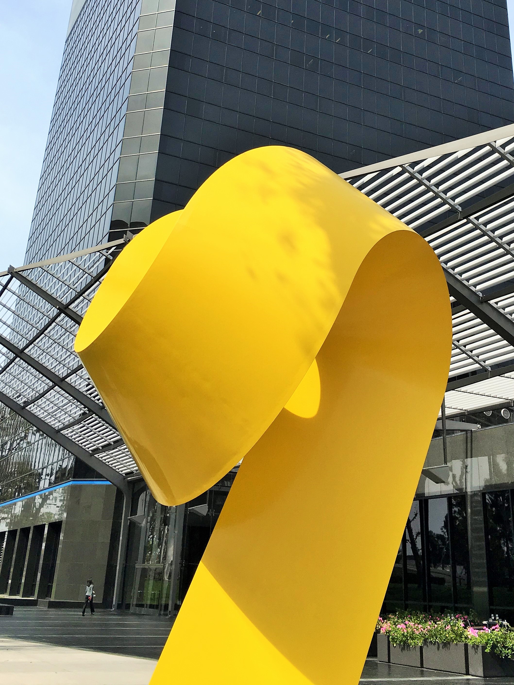 A large, bright yellow abstract sculpture stands in front of a modern glass office building in Costa Mesa, City of the Arts, with metal awnings and manicured landscaping on a sunny day.