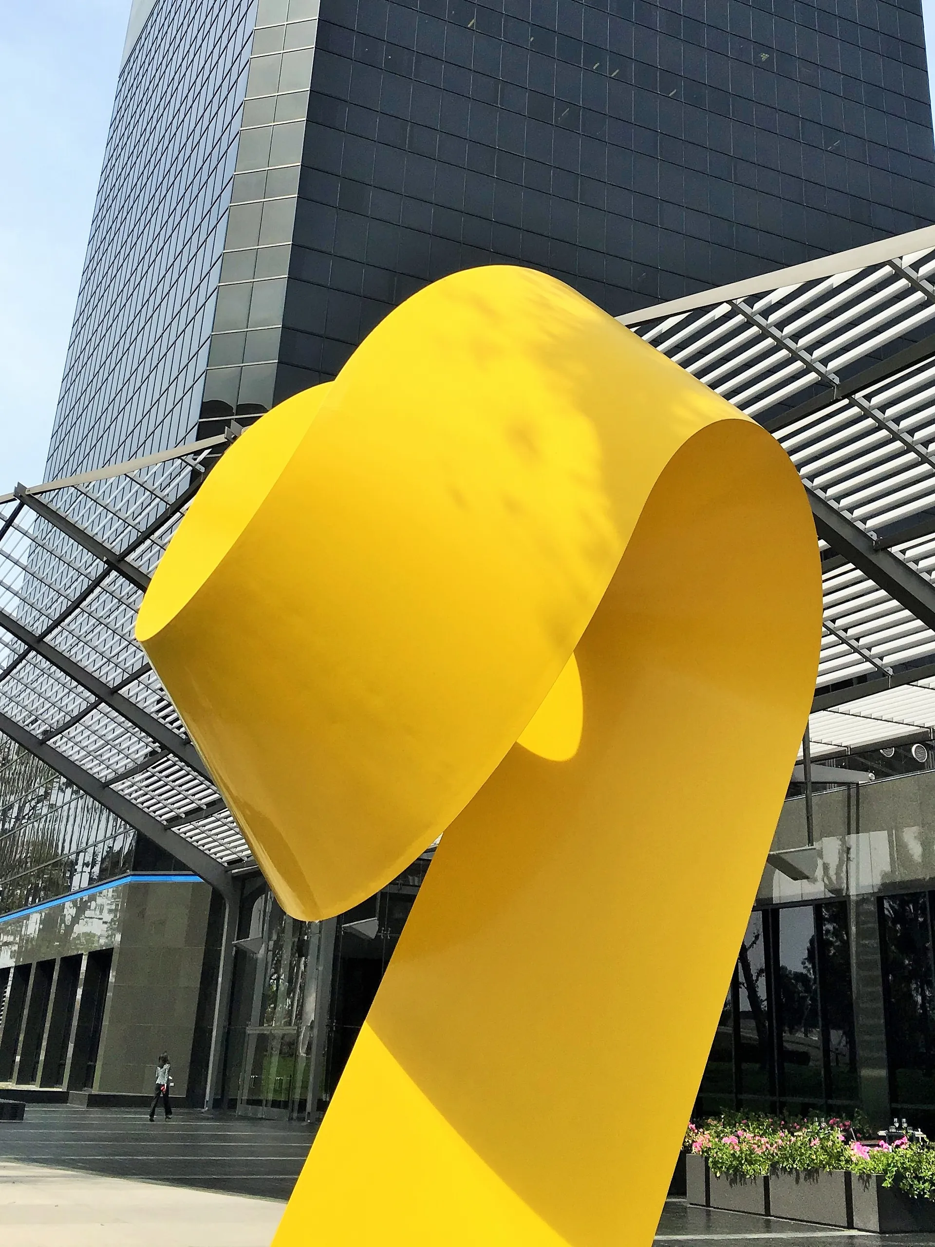 A large, bright yellow abstract sculpture stands in front of a modern glass office building in Costa Mesa, City of the Arts, with metal awnings and manicured landscaping on a sunny day.