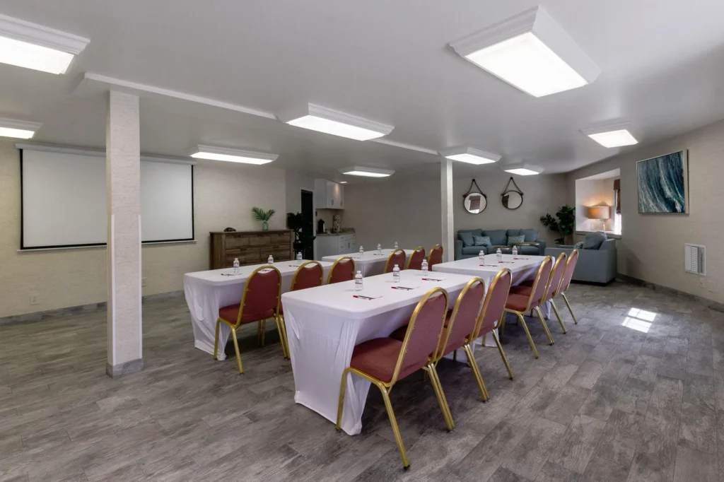 A conference room at one of the top event venues in Costa Mesa, featuring two tables draped in white cloths, red chairs, water bottles, notepads, a projector screen, and a cozy seating area with sofas and stylish decor.