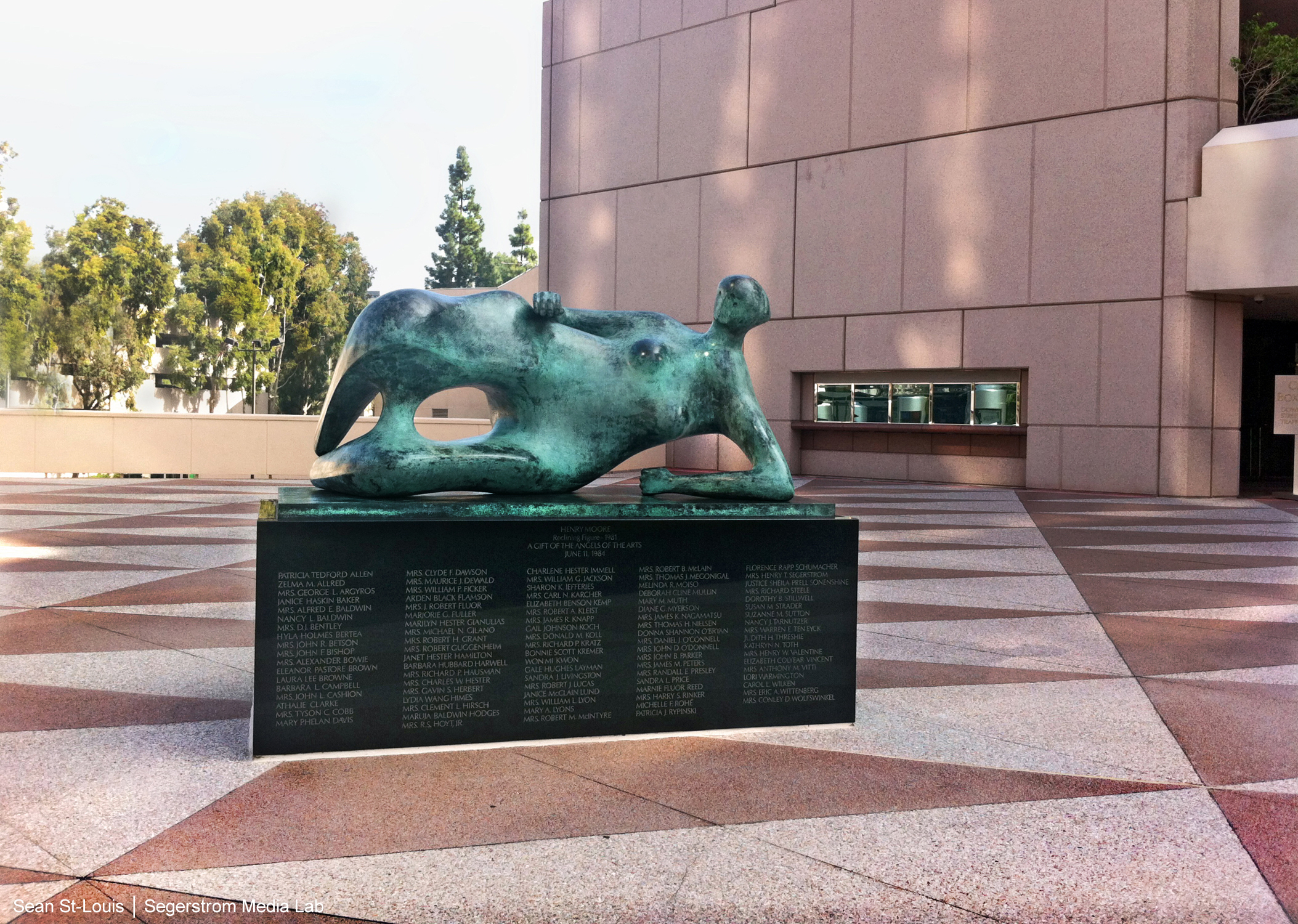 A modern abstract bronze sculpture of a reclining human figure rests on a black pedestal inscribed with names, situated in a spacious outdoor plaza along the Costa Mesa Art Walk near a large tan building and trees.