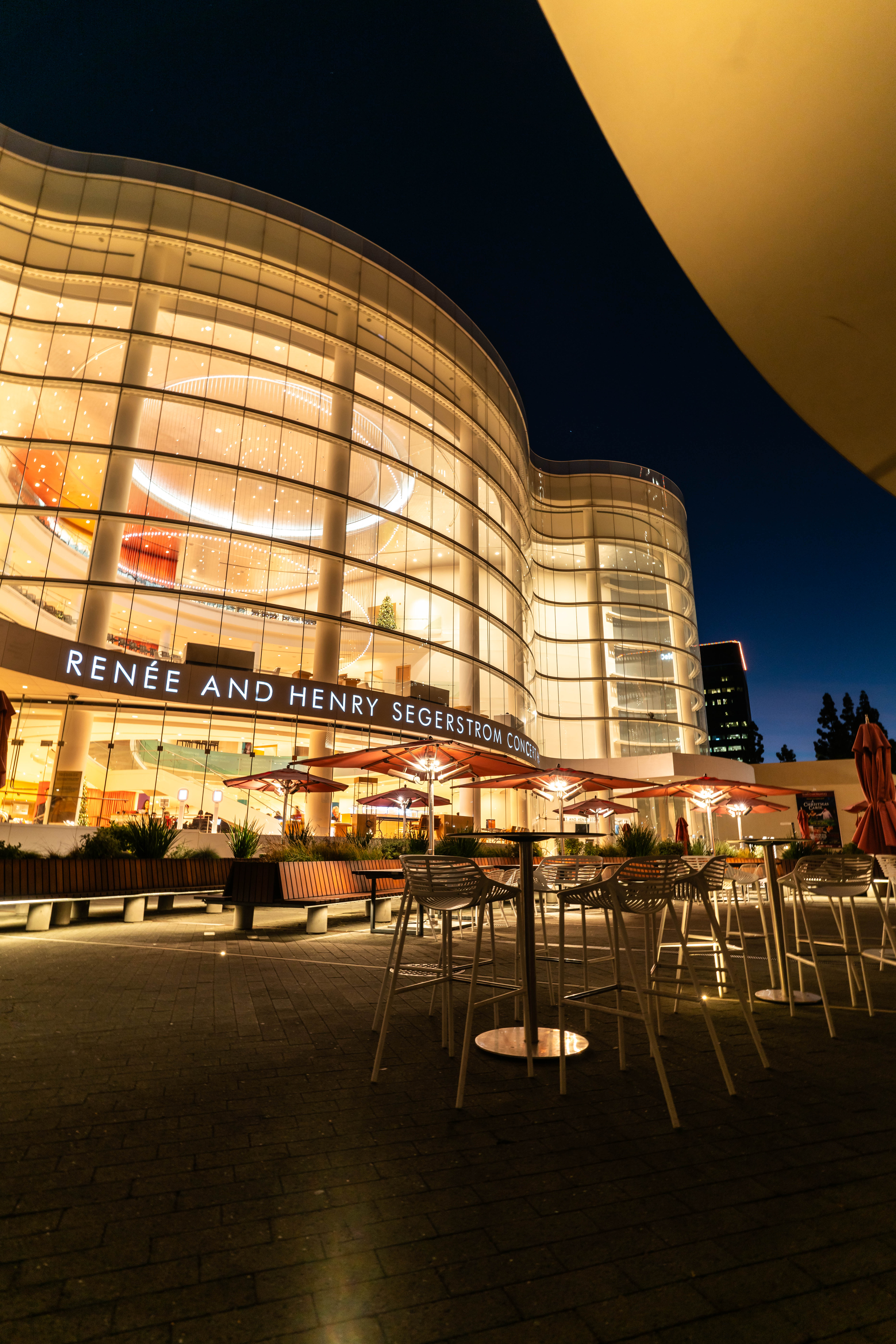 Outdoor seating area at night in front of the illuminated, modern glass facade of the Renée and Henry Segerstrom Concert Hall, highlighting Costa Mesa City of the Arts​, with empty tables and chairs under soft lighting.
