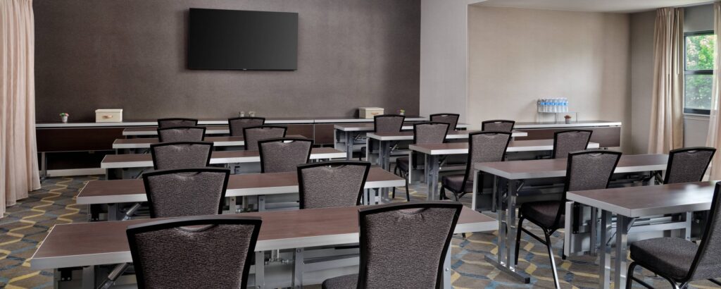 A modern classroom with rows of empty desks and chairs facing a large wall-mounted TV, similar to top event venues in Costa Mesa. Sunlight enters through windows on the right, highlighting neutral colors and a water dispenser in the corner.