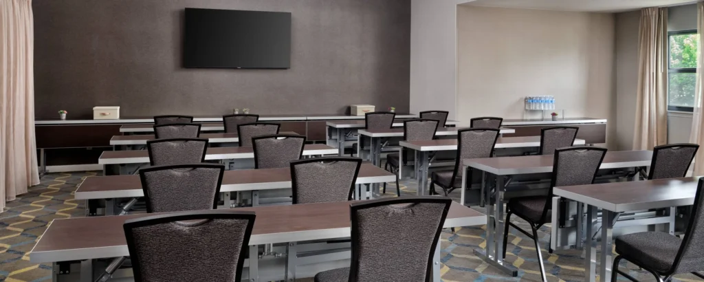 A modern classroom with rows of empty desks and chairs facing a large wall-mounted TV, similar to top event venues in Costa Mesa. Sunlight enters through windows on the right, highlighting neutral colors and a water dispenser in the corner.