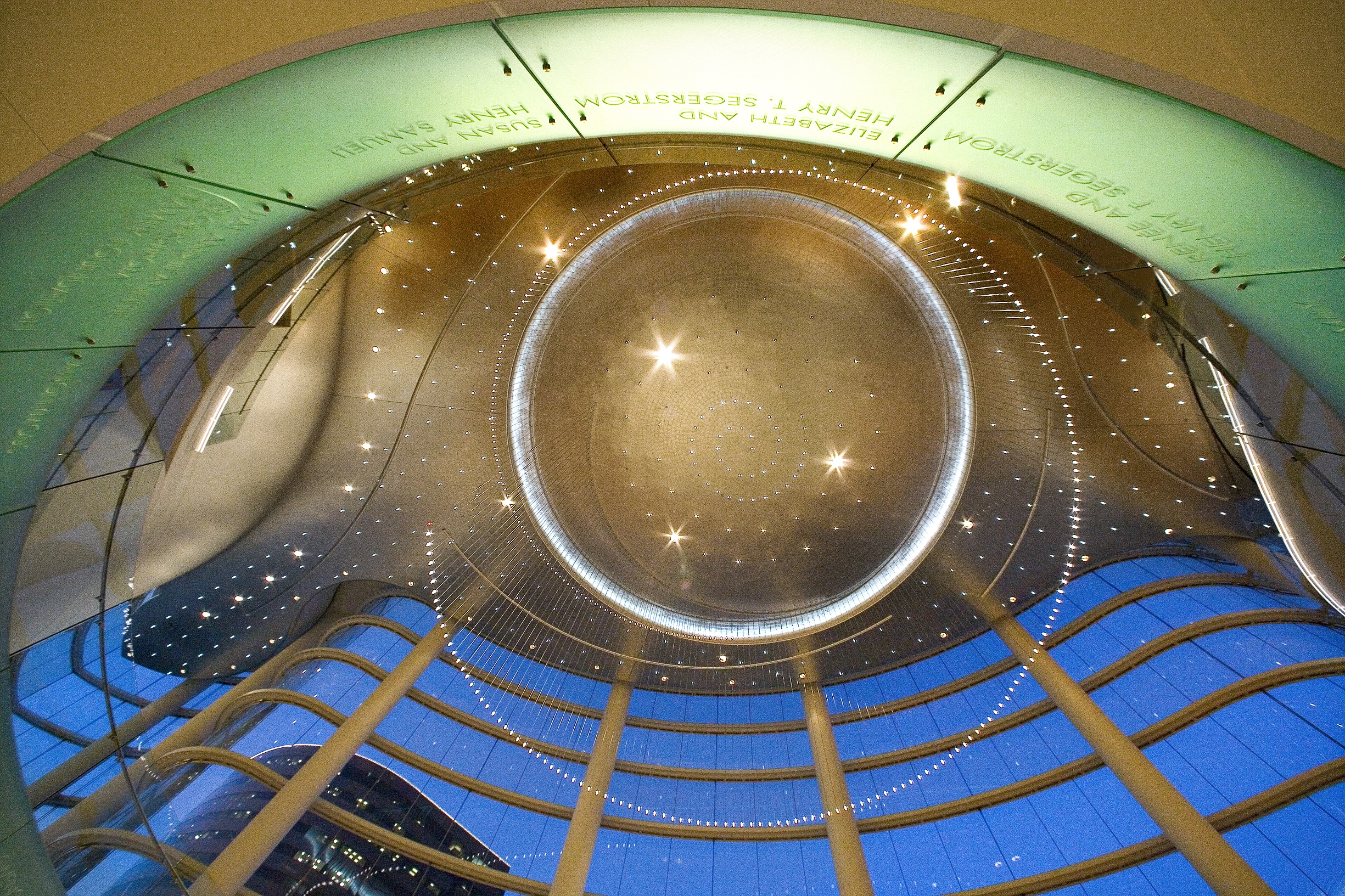 Interior view looking up at a modern glass and steel atrium ceiling, ringed with lights and reflections, with tall columns and curved glass panels at dusk—a scene reminiscent of the Costa Mesa Art Walk. Names are etched on a green circular panel above.