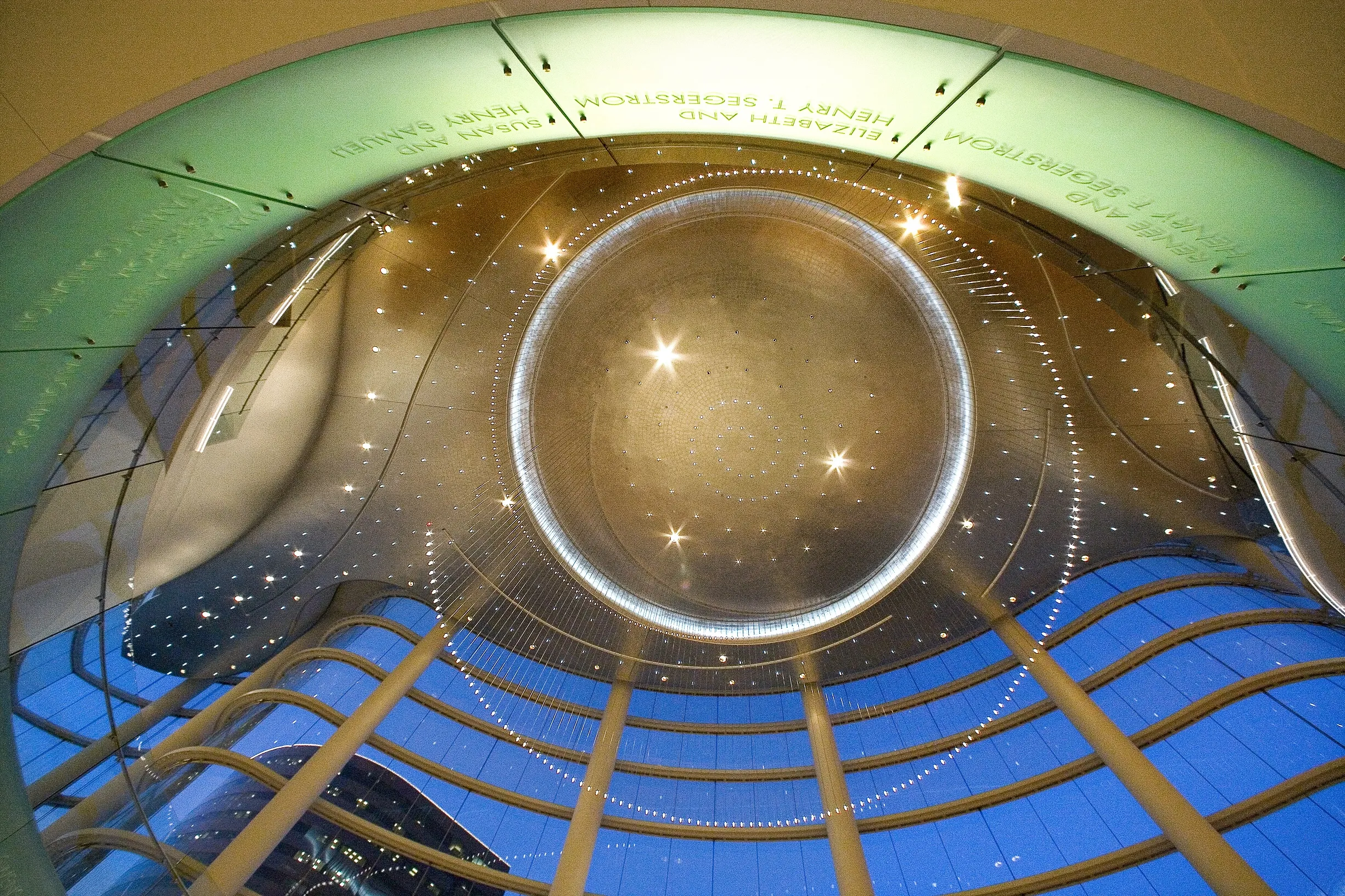 Interior view looking up at a modern glass and steel atrium ceiling, ringed with lights and reflections, with tall columns and curved glass panels at dusk—a scene reminiscent of the Costa Mesa Art Walk. Names are etched on a green circular panel above.