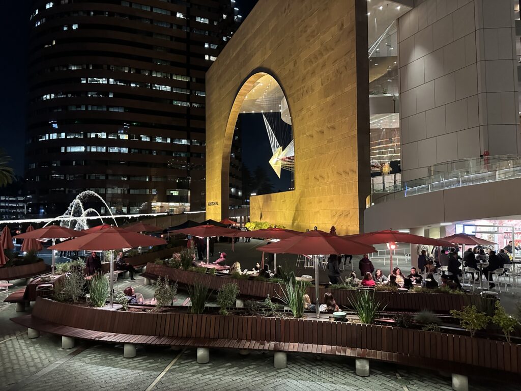 Outdoor plaza at night with people sitting under red umbrellas, a modern building with a large arch illuminated in yellow—one of the vibrant event venues in Costa Mesa—plus a water fountain to the left and city lights glowing in the background.