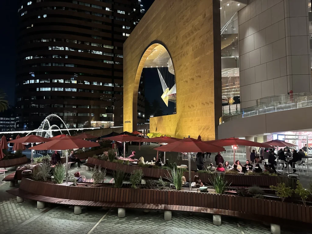Outdoor plaza at night with people sitting under red umbrellas, a modern building with a large arch illuminated in yellow—one of the vibrant event venues in Costa Mesa—plus a water fountain to the left and city lights glowing in the background.