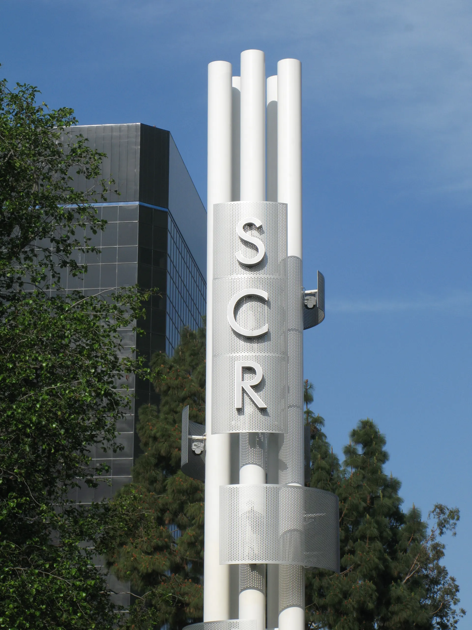 A tall white metal sculpture with the letters SCR stands in front of trees and a modern glass building under a blue sky at the Segerstrom Performing Arts Center in Costa Mesa.