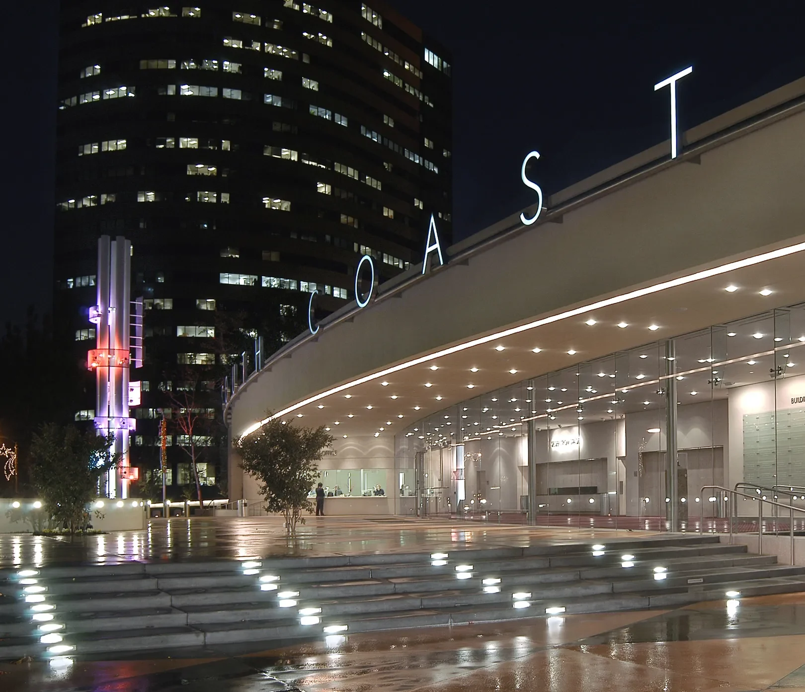 A modern, glass-walled building at night with COAST in large illuminated letters on the curved roof, bright outdoor lighting, and a wet reflective plaza in front, set against tall city buildings in Costa Mesa City of the Arts.