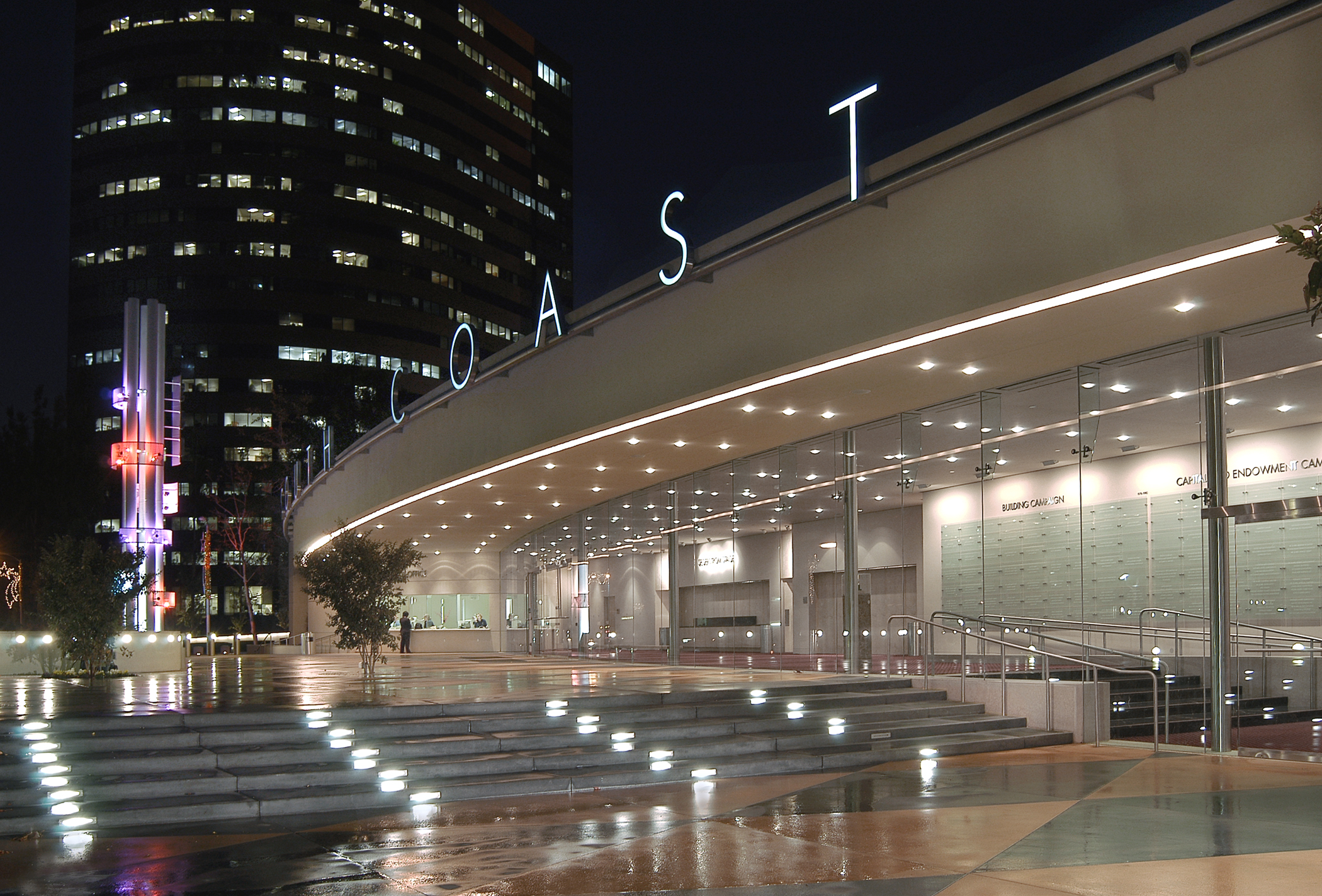 A modern, glass-walled building at night with COAST in large illuminated letters on the curved roof, bright outdoor lighting, and a wet reflective plaza in front, set against tall city buildings in Costa Mesa City of the Arts.