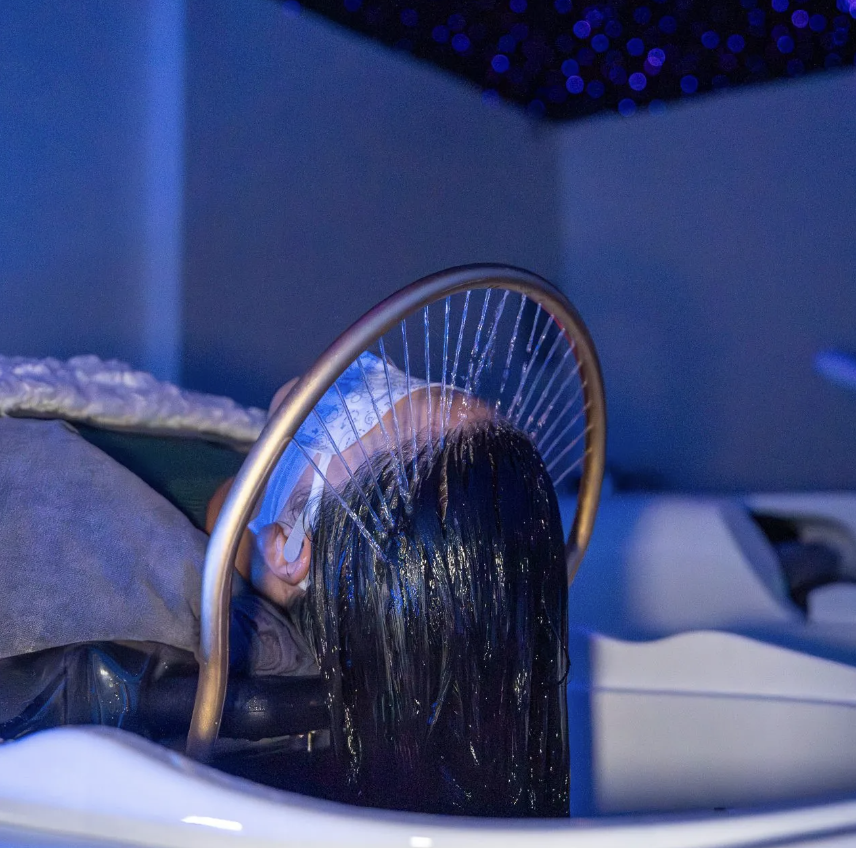 A person reclines at a salon sink with water streaming from a circular shower head onto their hair, under blue ambient lighting and a blanket draped over their shoulders—a relaxing treat among the top things to do in Costa Mesa.