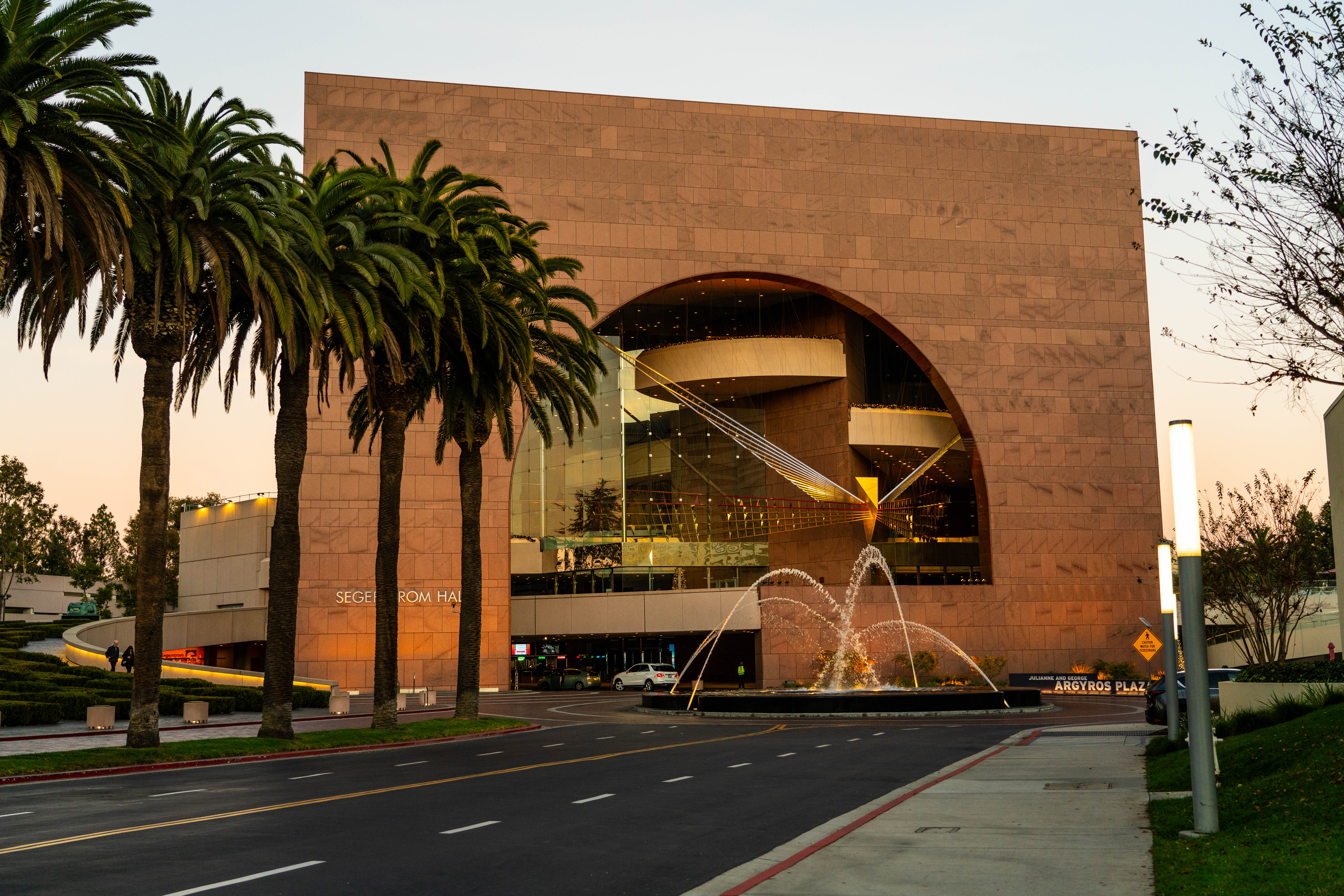 The image shows Segerstrom Hall in Costa Mesa, City of the Arts—a modern building with a large glass arch, palm trees, and a fountain in front at sunset. The street and sidewalk lead up to the entrance.